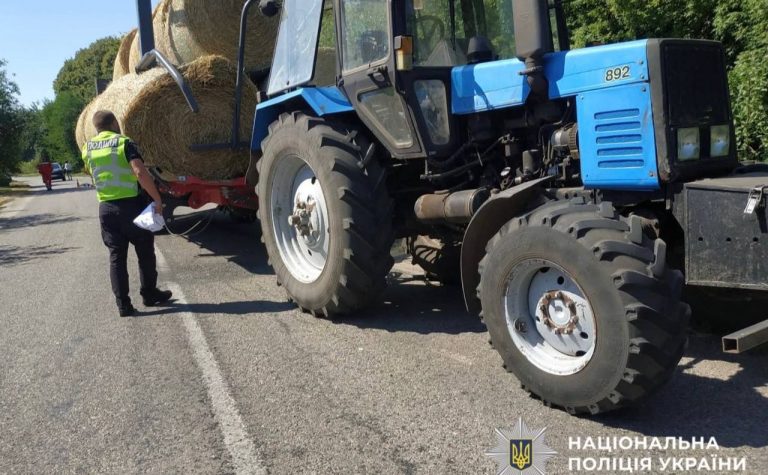 Поліцейський оглядає синій трактор із причепом, завантаженим тюками сіна.