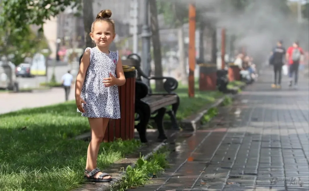 Маленька дівчинка стоїть на алеї в одному з парків Києва поруч із лавками та системою водяного охолодження в спекотний літній день.